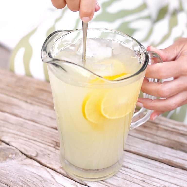 Jar of lemonade with lemon slices being stirred on a light wooden table.