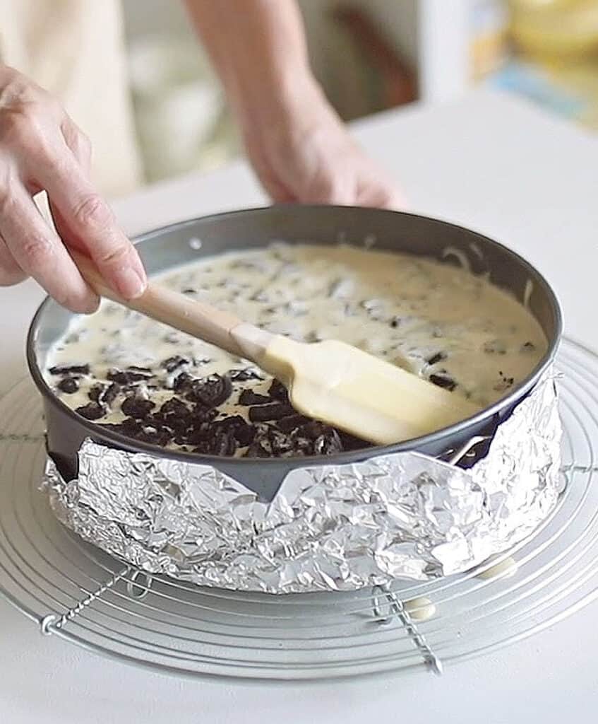 Submerging oreo pieces on cheesecake batter with a yellow spatula. White surface.