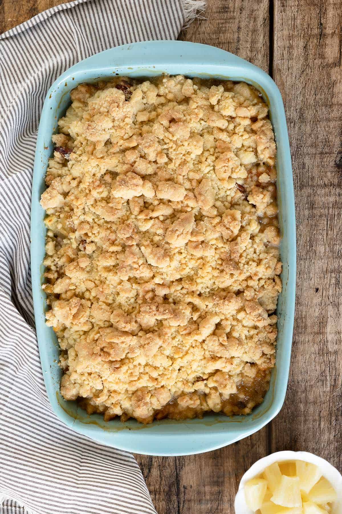 Baked pineapple dump cake in a rectangular blue baking dish. Wooden surface and striped cloth.