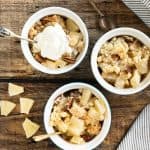 Servings of pineapple dump cake in three white bowls on a wooden surface. Spoons, a striped cloth, and chunks of pineapple around.