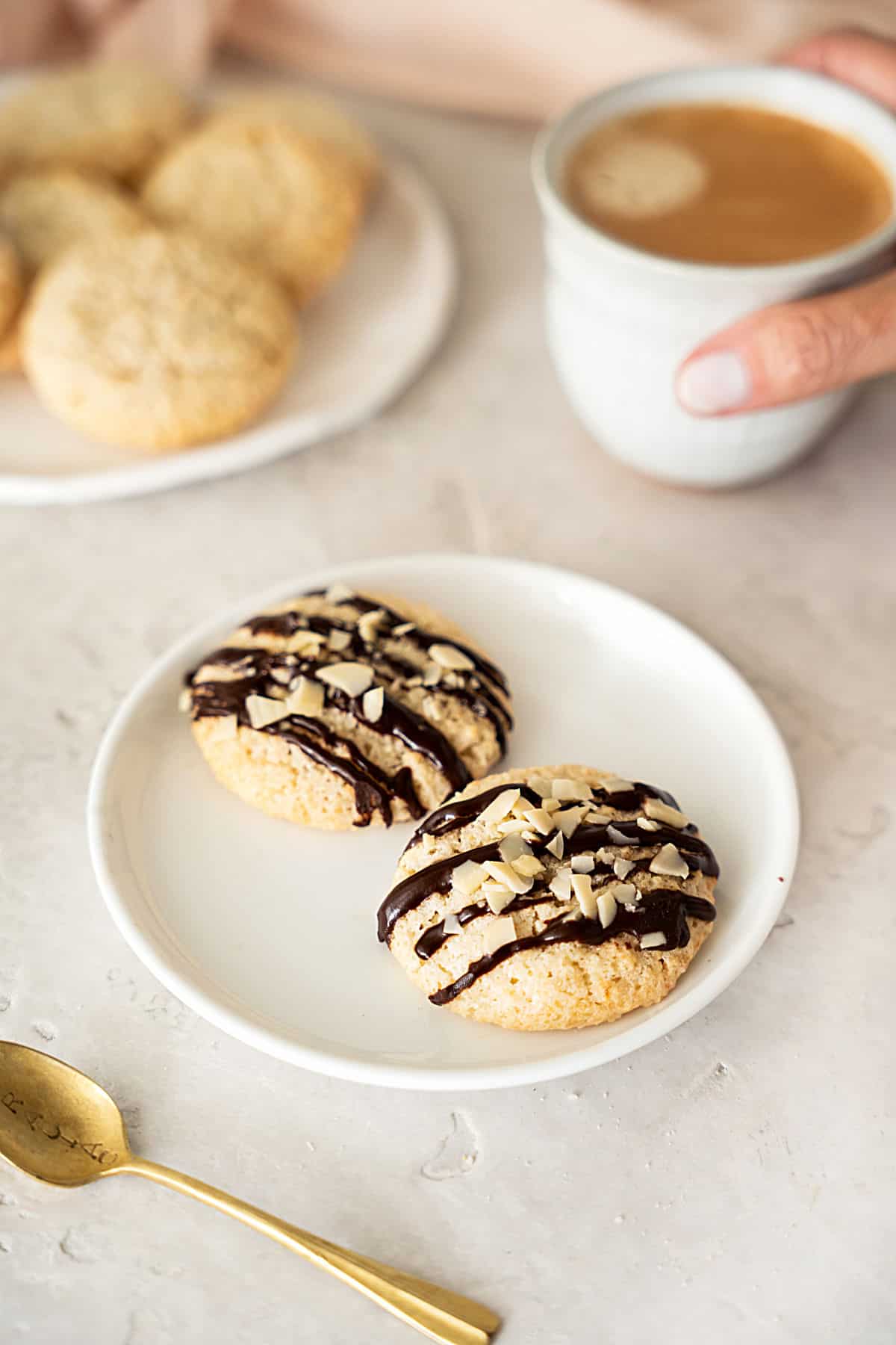 White plates with plain and chocolate drizzled almond cookies. Hand holding coffee cup, golden spoon, grey surface.
