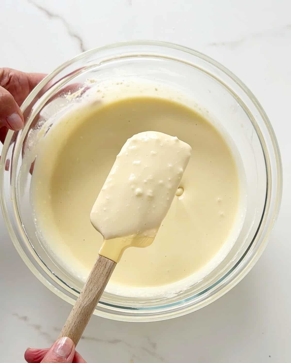 Ricotta cheesecake batter texture being shown with a spatula from a glass bowl. White marble surface.