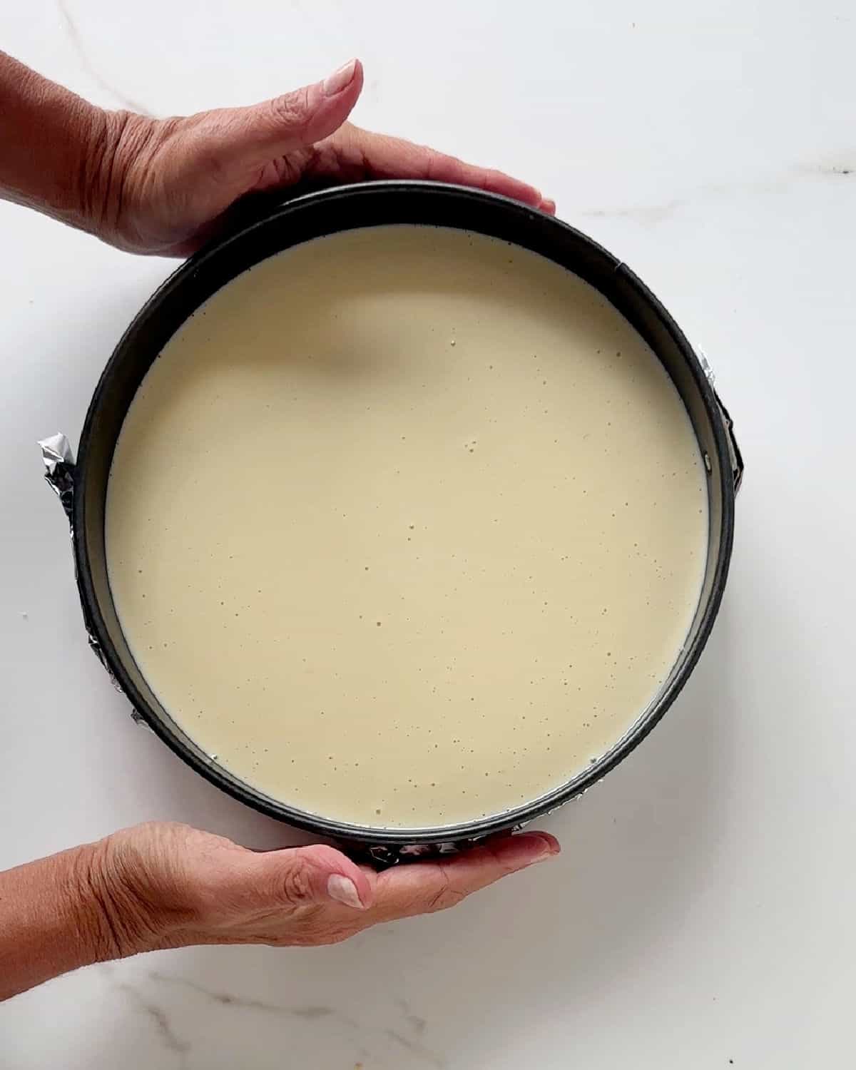 Hands holding a round cake pan with ricotta cheesecake filling before baking. White marble surface. 