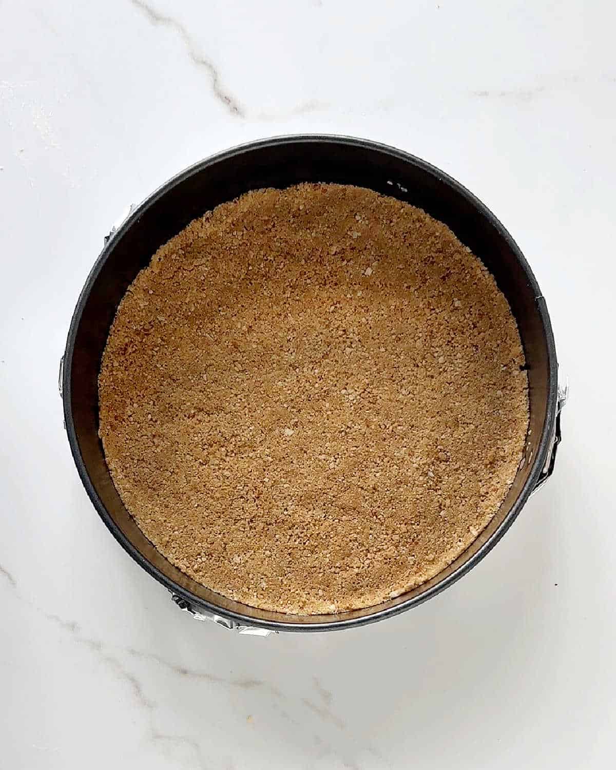 Graham cracker crust for cheesecake in a round dark metal cake pan on white marble.
