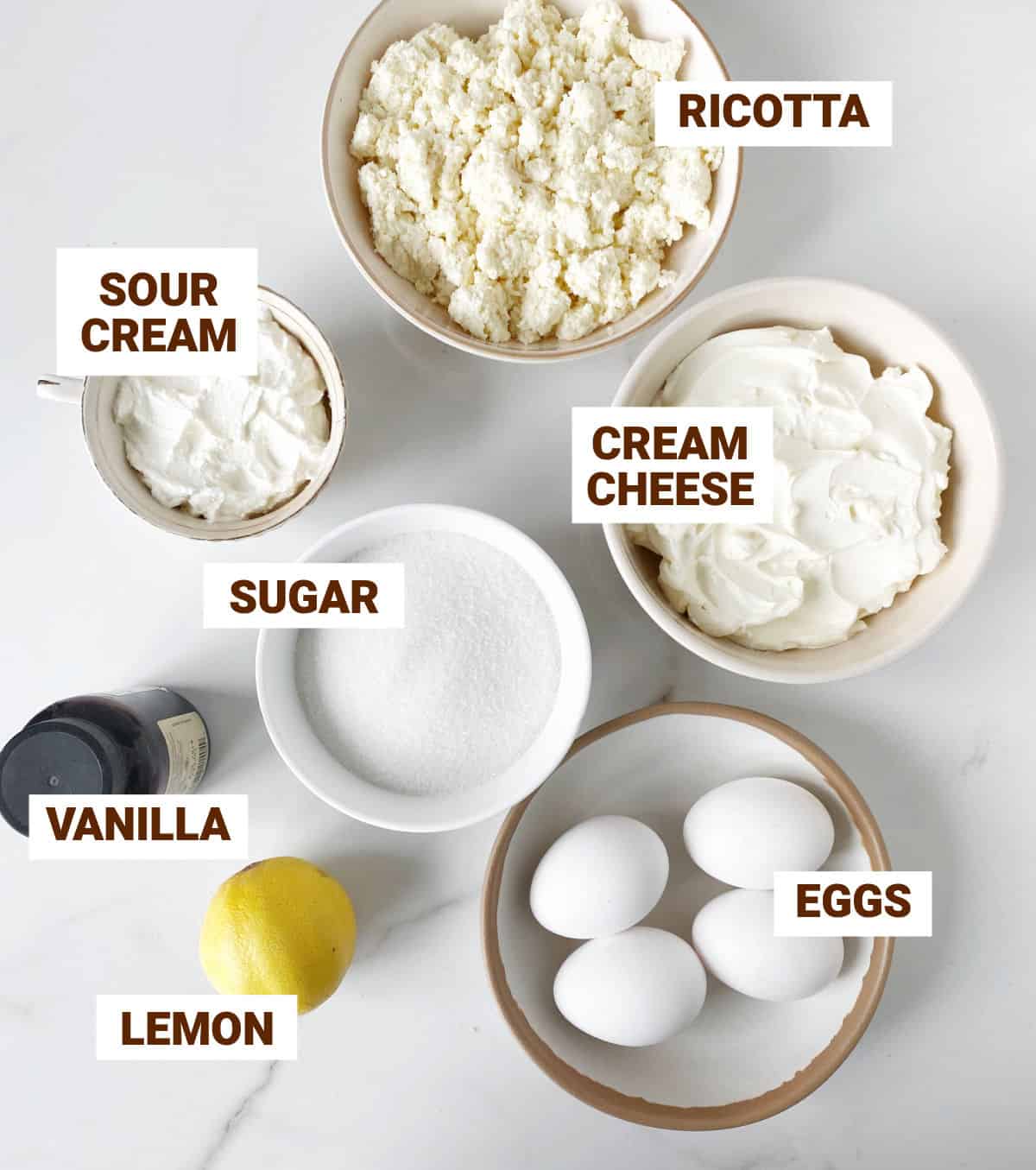 Ingredients for ricotta cheesecake in bowls on white marble, including lemon, vanilla, sugar, eggs, sour cream, cream cheese.
