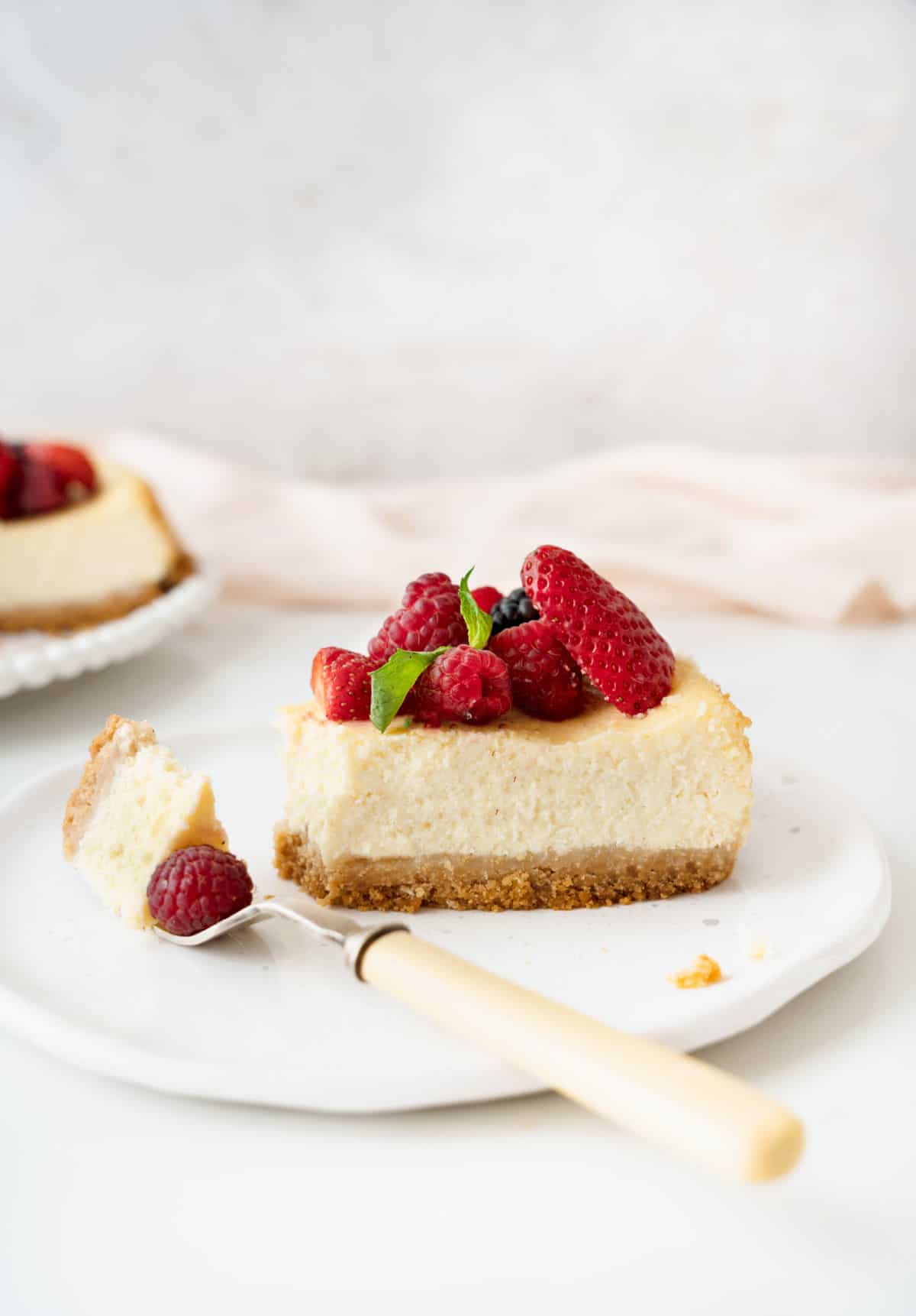 Slice of ricotta berry cheesecake on a white plate with bite in a fork. Light grey, pink, and white surface and background. 