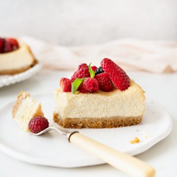 Single slice of ricotta cheesecake with berries on a white plate with a fork. Light pink cloth on a grey background.