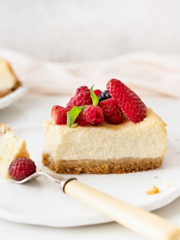 Single slice of ricotta cheesecake with berries on a white plate with a fork. Light pink cloth on a grey background.