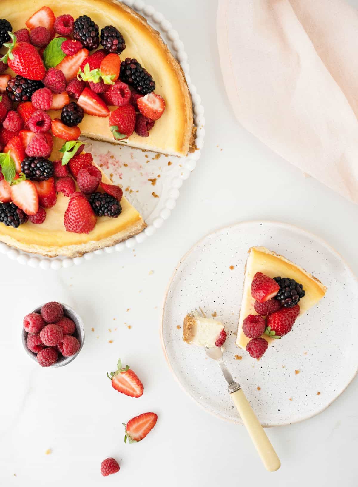 Top view of ricotta berry cheesecake on a plate with a slice missing that's on a white plate with a fork. White surface.