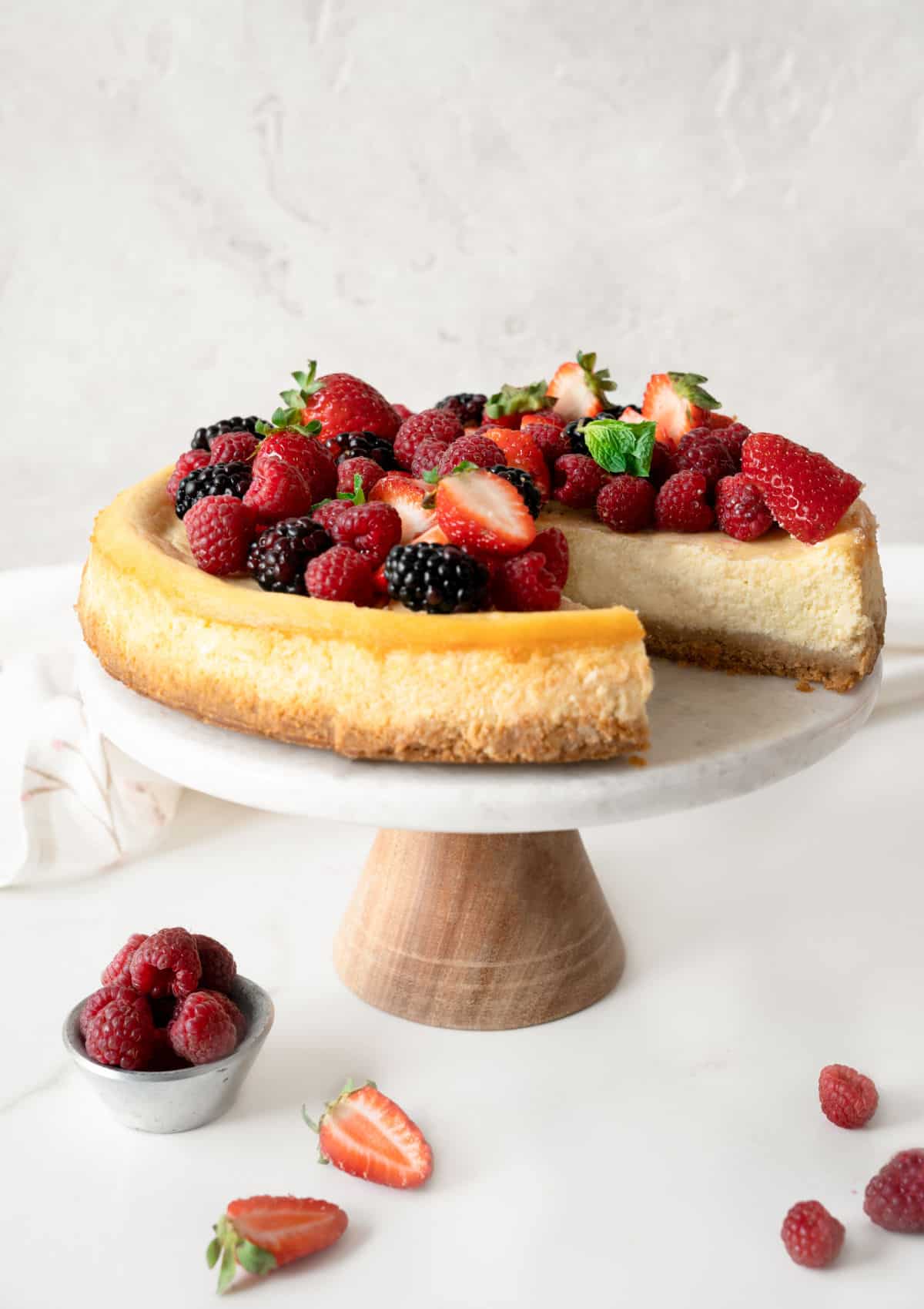Wooden and white marble cake stand with berry ricotta cheesecake with a missing slice. Grey background, white surface and loose berries. 