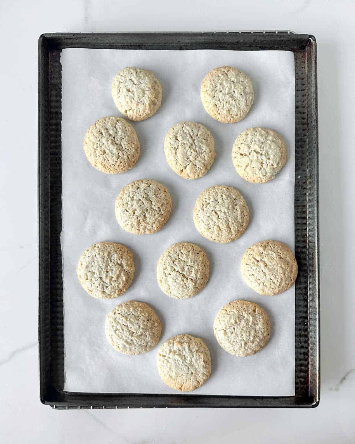 Baked almond macaroons on white parchment paper on a dark metal cookie sheet. White marble surface.