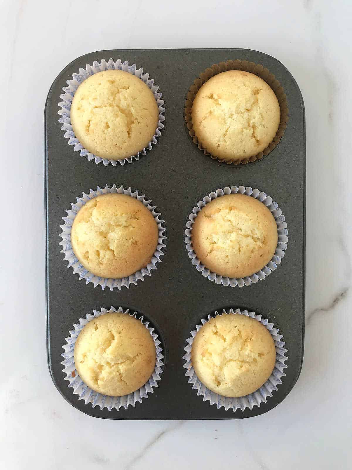 Six baked vanilla cupcakes in a metal muffin pan. White marbled surface.