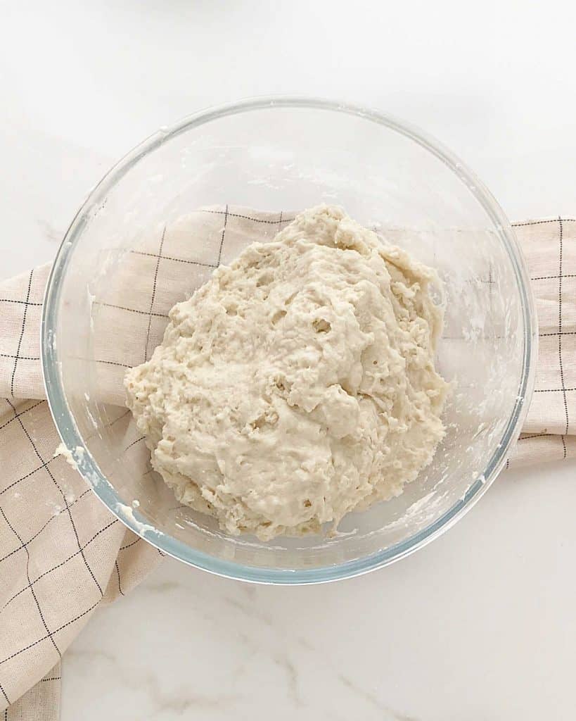 Glass bowl with beer bread batter. White marble surface with beige kitchen towel.