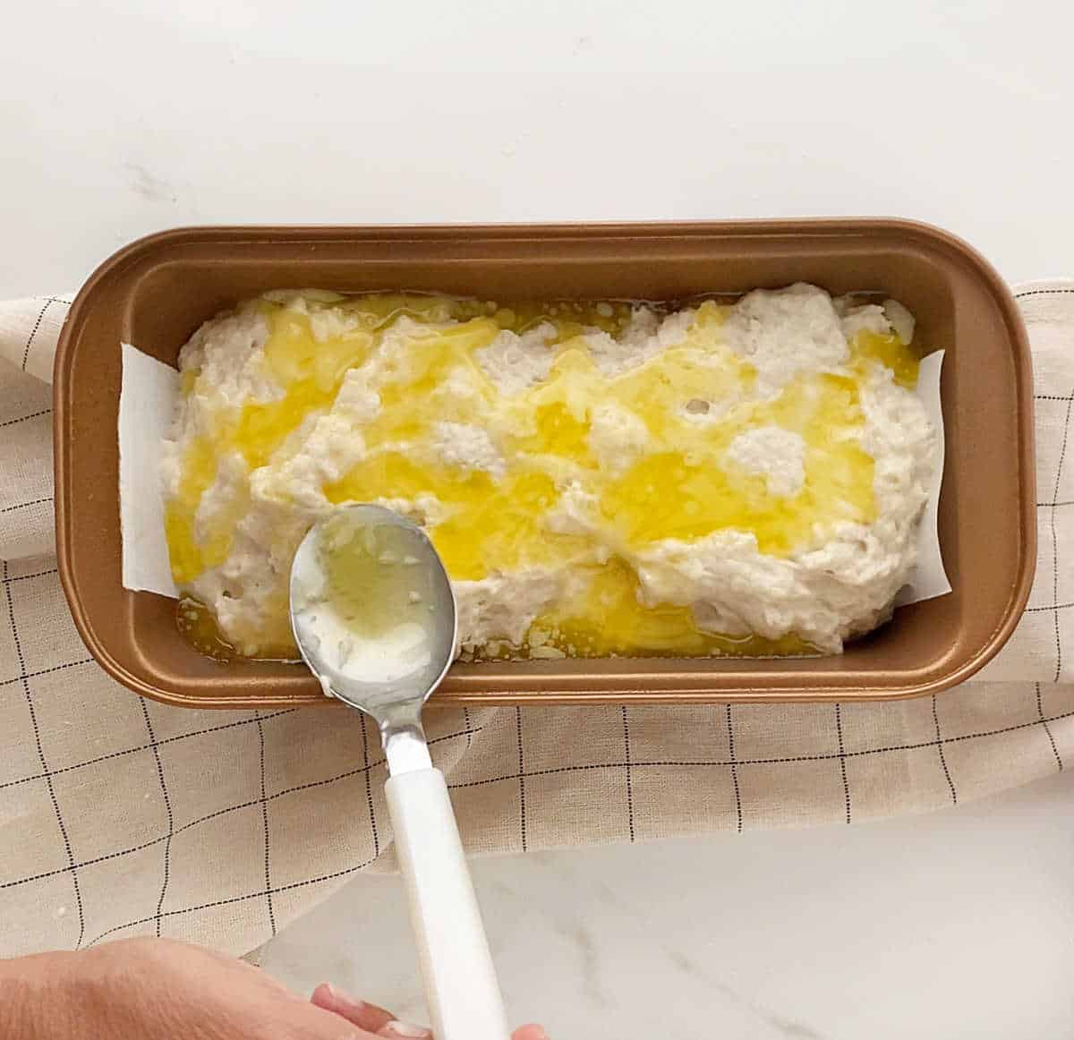 Melted butter being added with a spoon to beer bread batter in a copper loaf pan set on a beige towel. White marble surface.
