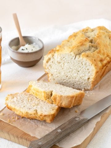 Close up of beer bread loaf with slices on a wooden board. Glass of beer, knife, small bowl, white pink background.