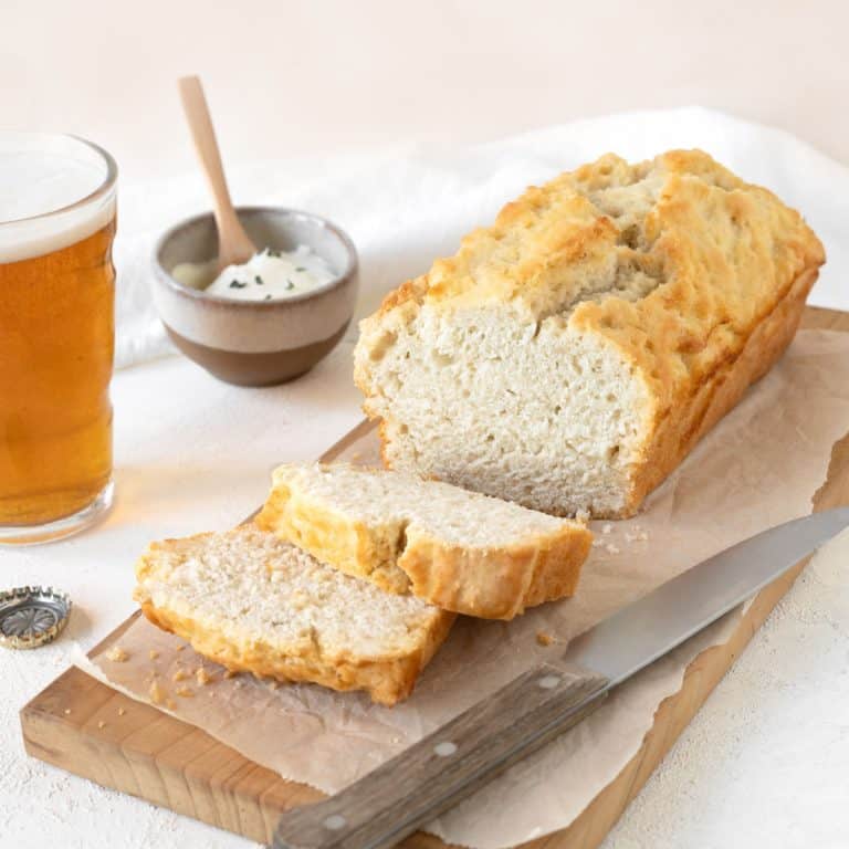 Close up of beer bread loaf with slices on a wooden board. Glass of beer, knife, small bowl, white pink background.