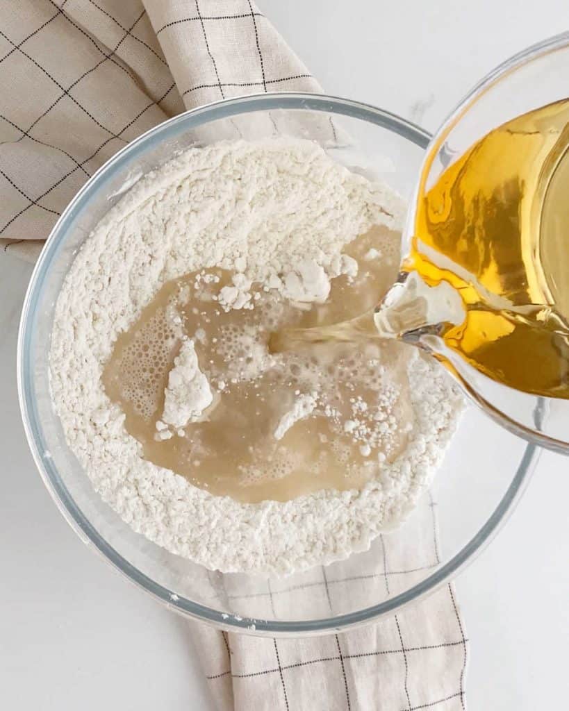 Adding beer to a glass bowl with flour mixture on a checkered beige cloth on a white marble surface.