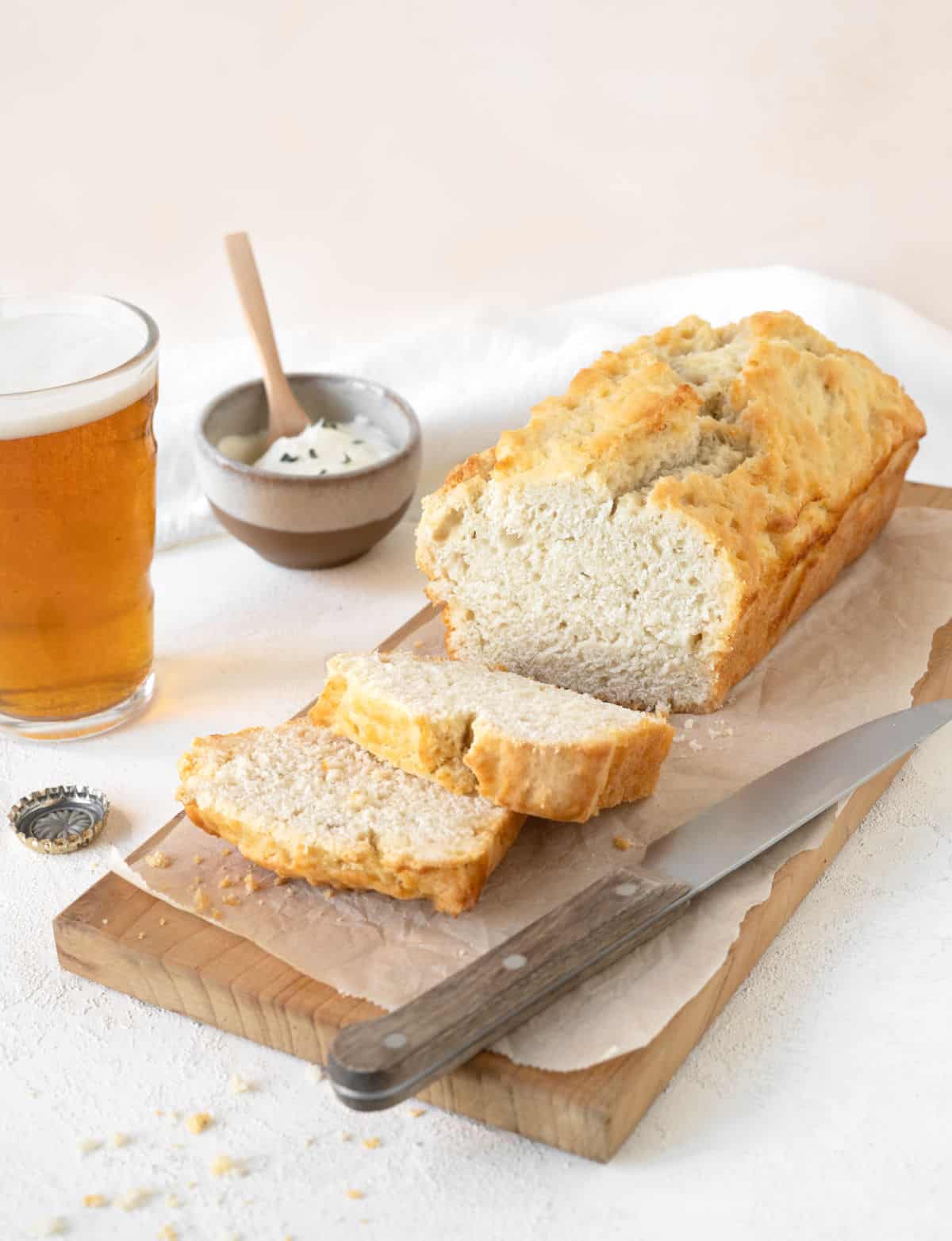 Wooden board with parchment paper on white marble with loaf of beer bread, whole and sliced. A knife, glass of beer and pinkish background. 