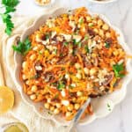 Top view of round white bowl with chickpea carrot dried fruit salad. Silver spoon. White surface and cloth.