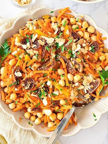 Top view of round white bowl with chickpea carrot dried fruit salad. Silver spoon. White surface and cloth.