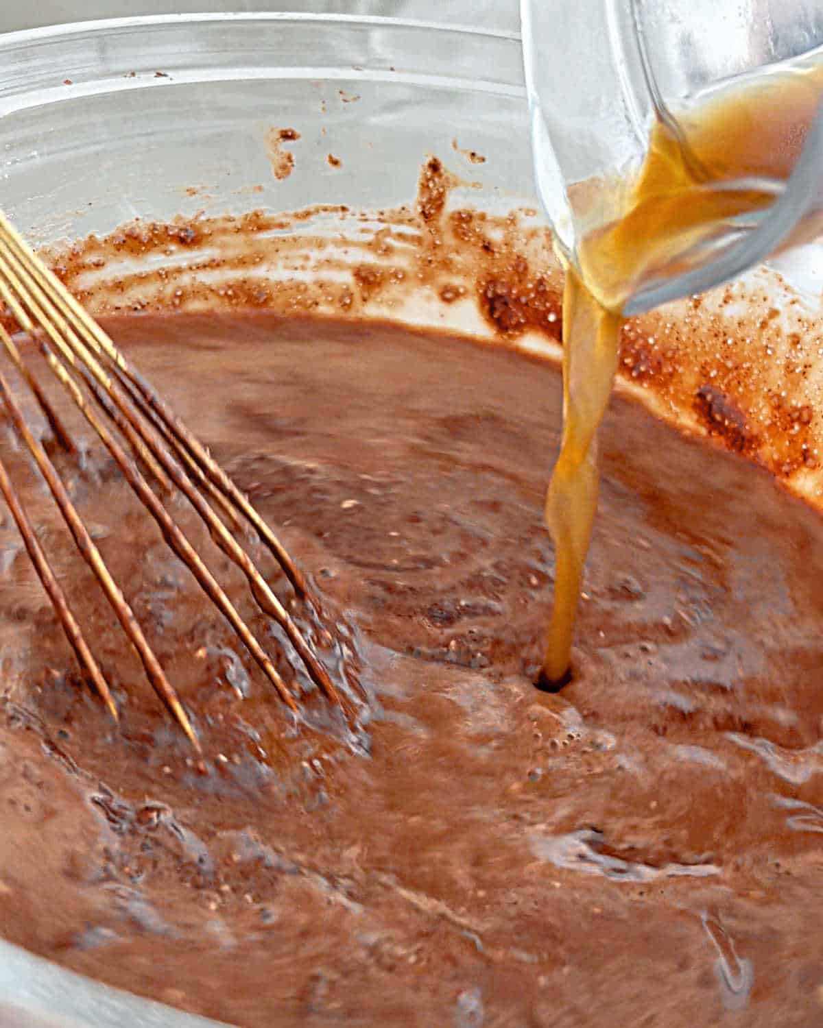 Adding coffee to a chocolate cake batter in a glass bowl with a whisk inside.