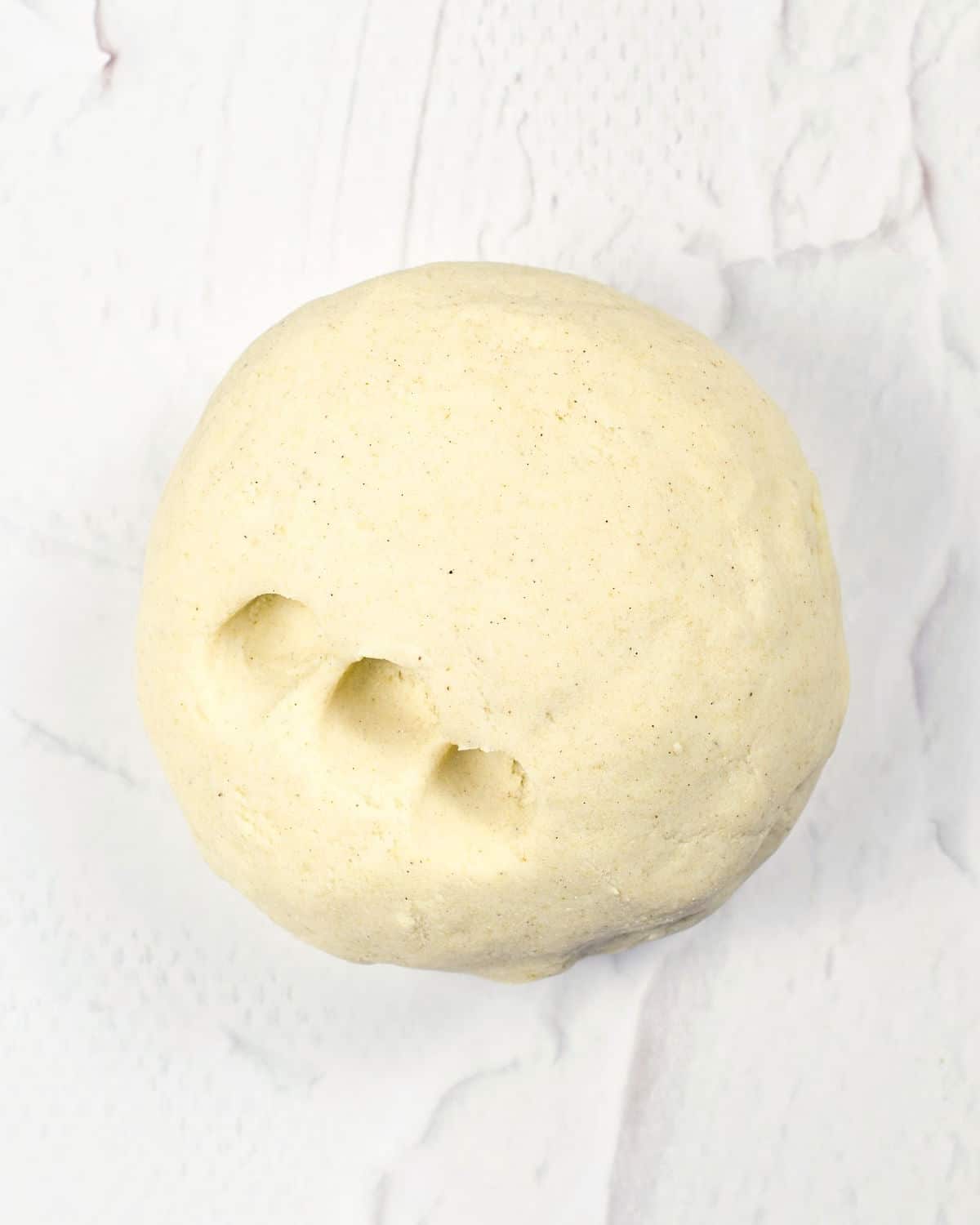 Ball of tortilla dough with three indentations on a textured white greyish surface.