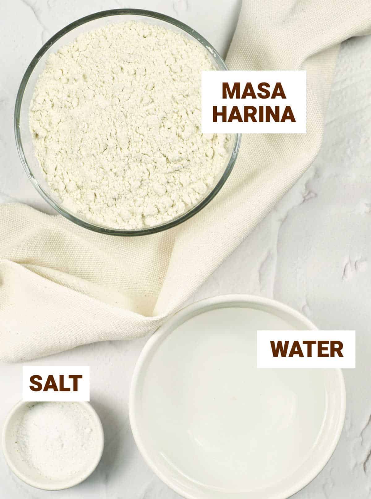White surface with a kitchen towel and bowls containing masa harina, salt, and water.