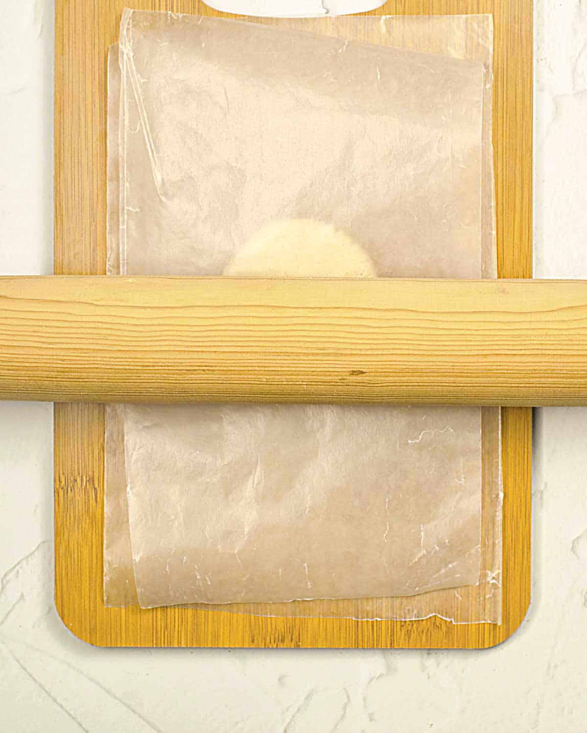 Rolling pin, a wooden board, and a tortilla dough ball between sheets of wax paper. Grey surface.
