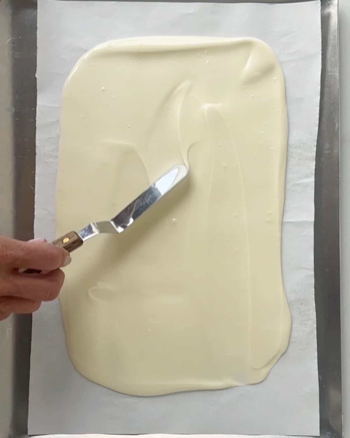 Spreading melted white chocolate with an offset spatula on white parchment paper.