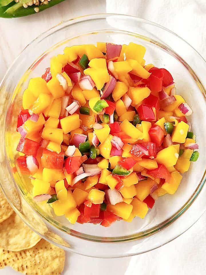 A glass bowl with mango peach salsa on a white surface with a white cloth and corn chips around.