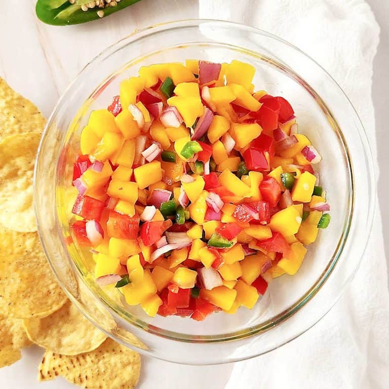 A glass bowl with mango peach salsa on a white surface with a white cloth and corn chips around.