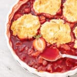 Close up of strawberry cobbler with silver spoon in a white baking dish on a grey surface.