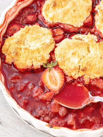 Close up of strawberry cobbler with silver spoon in a white baking dish on a grey surface.