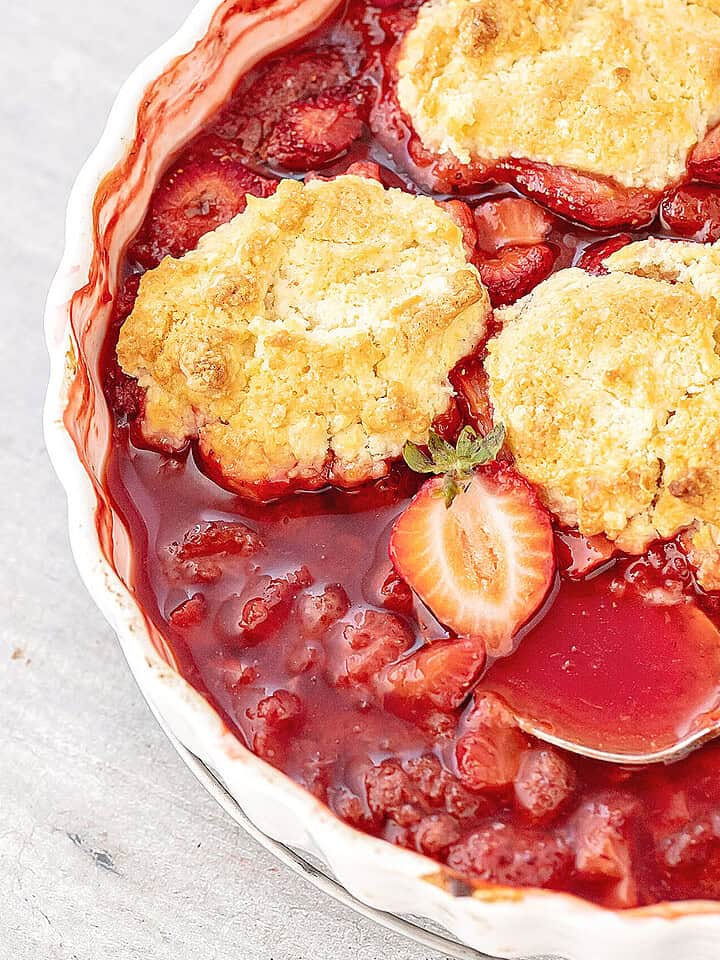 Close up of strawberry cobbler with silver spoon in a white baking dish on a grey surface.