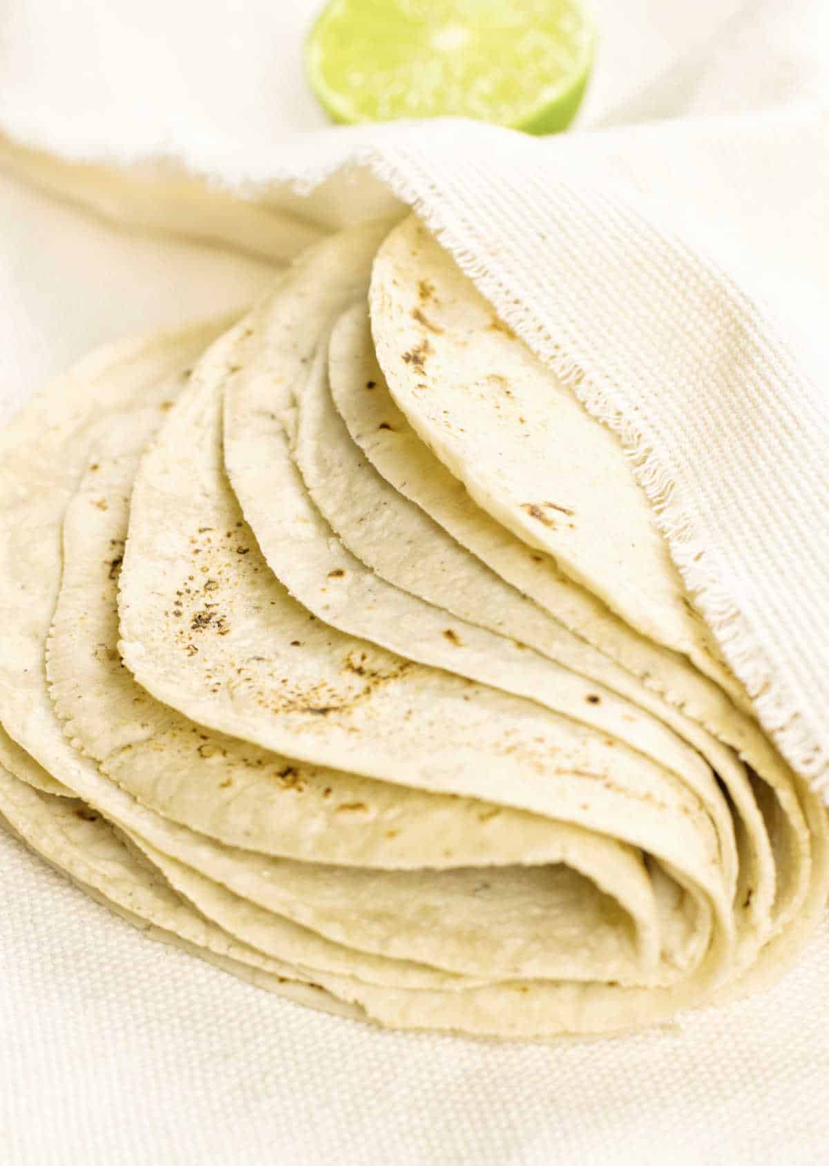 Folded stack of corn tortillas half covered by a cream colored cloth. Half a lime in the background. 