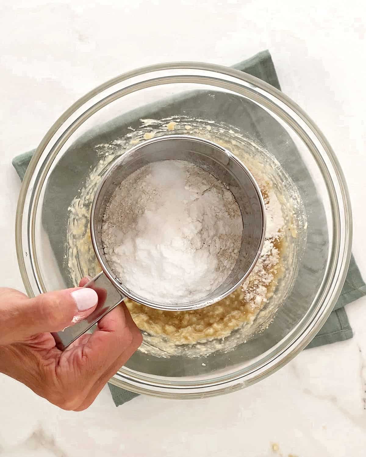 Sifting flour over banana muffin batter in a glass bowl. Green towel beneath it. White marble surface.