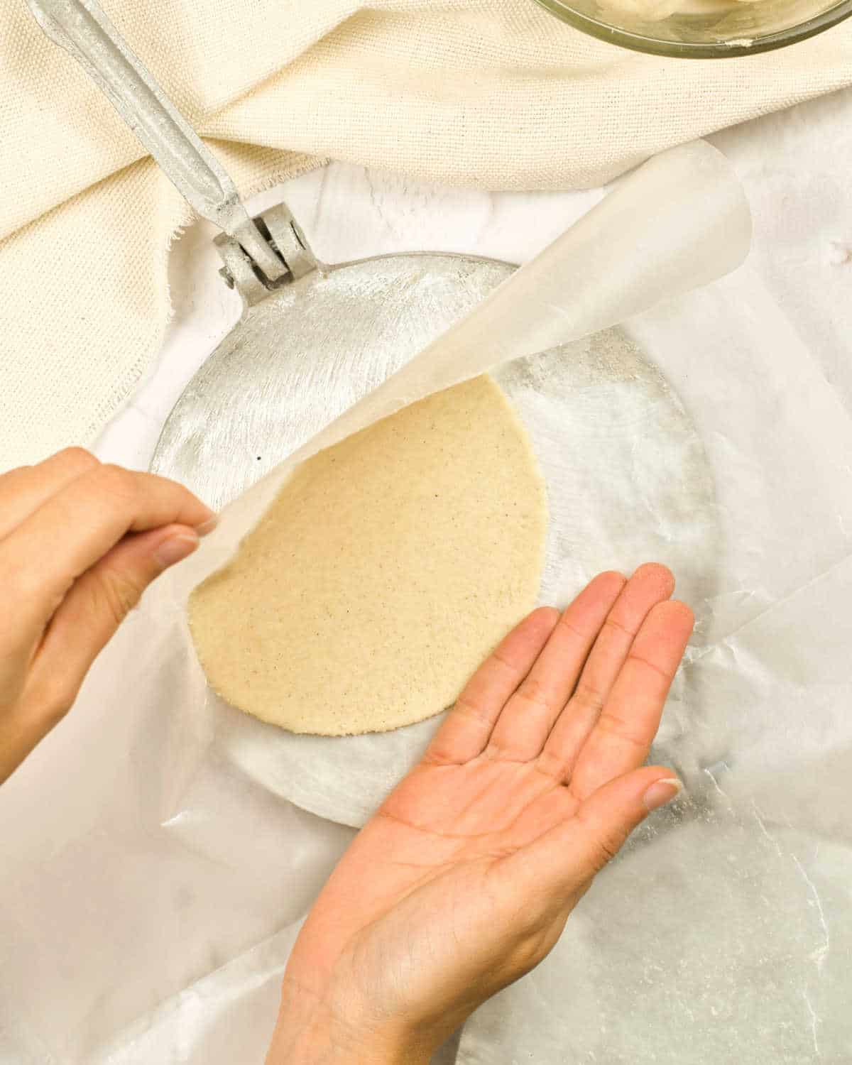 Lifting plastic sheet from a tortilla press to release the tortilla. Cream colored background.