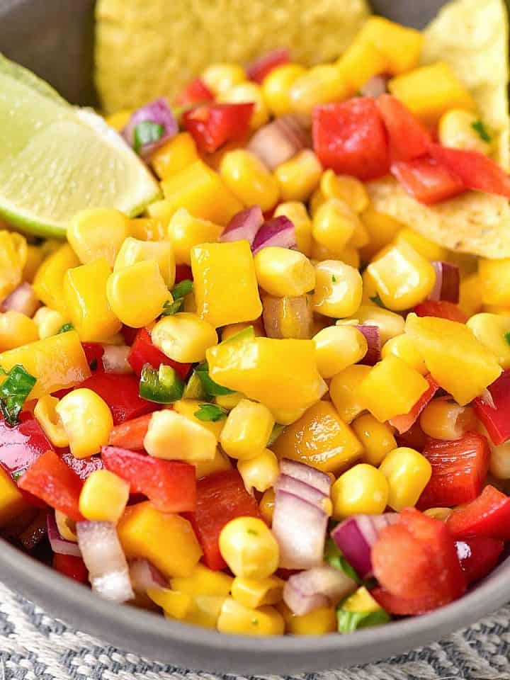 Close up of mango corn salsa in a grey bowl with lime wedge and chips.