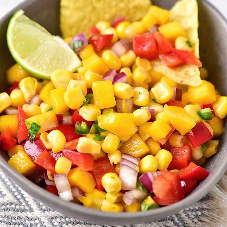 Close up of mango corn salsa in a grey bowl with lime wedge and chips.