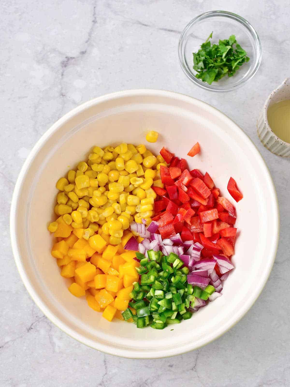 Ingredients for mango corn salsa in a white bowl and on a grey marble surface. 