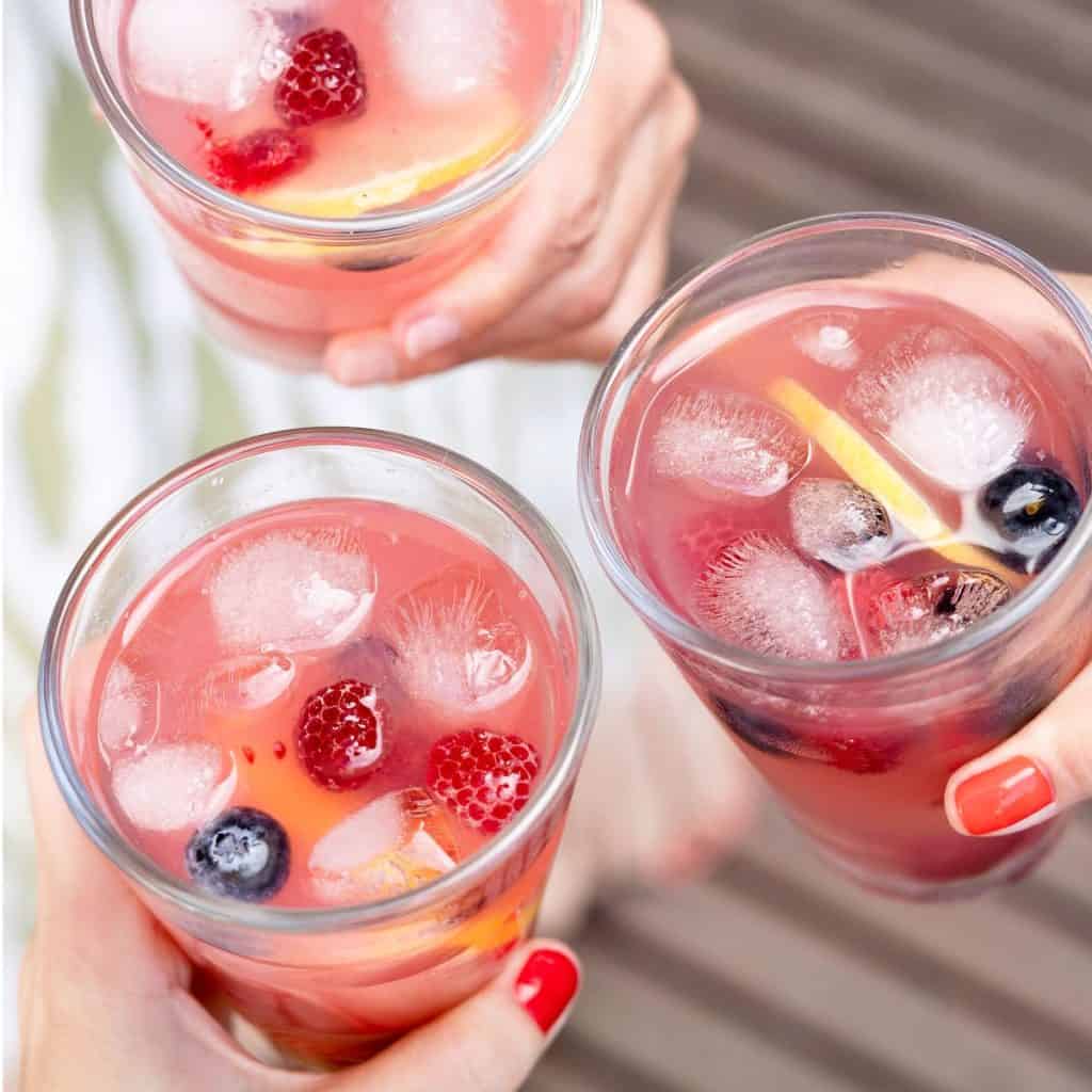 Hands holding three glasses with pink lemonade, ice cubes, and berries.