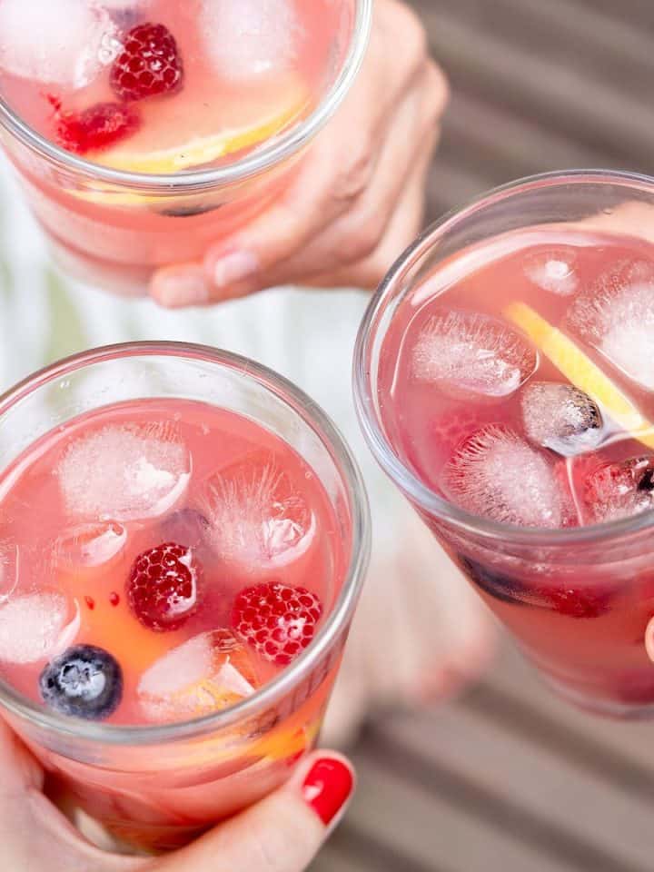 Hands holding three glasses with pink lemonade, ice cubes, and berries.