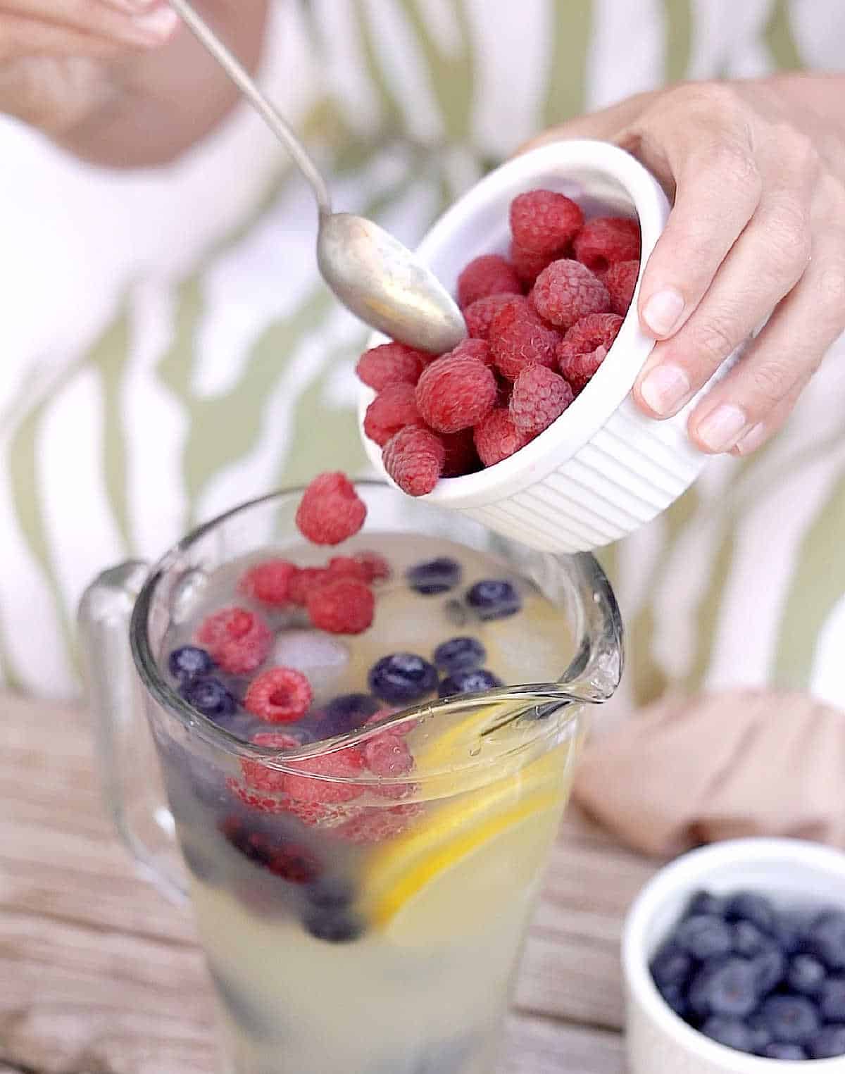 Adding fresh berries from a white to a lemonade pitcher. Green and white background.
