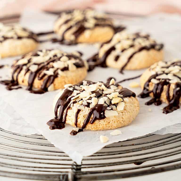 Piece of parchment paper with chocolate drizzled almond cookies on top of a wire rack. Pink background.