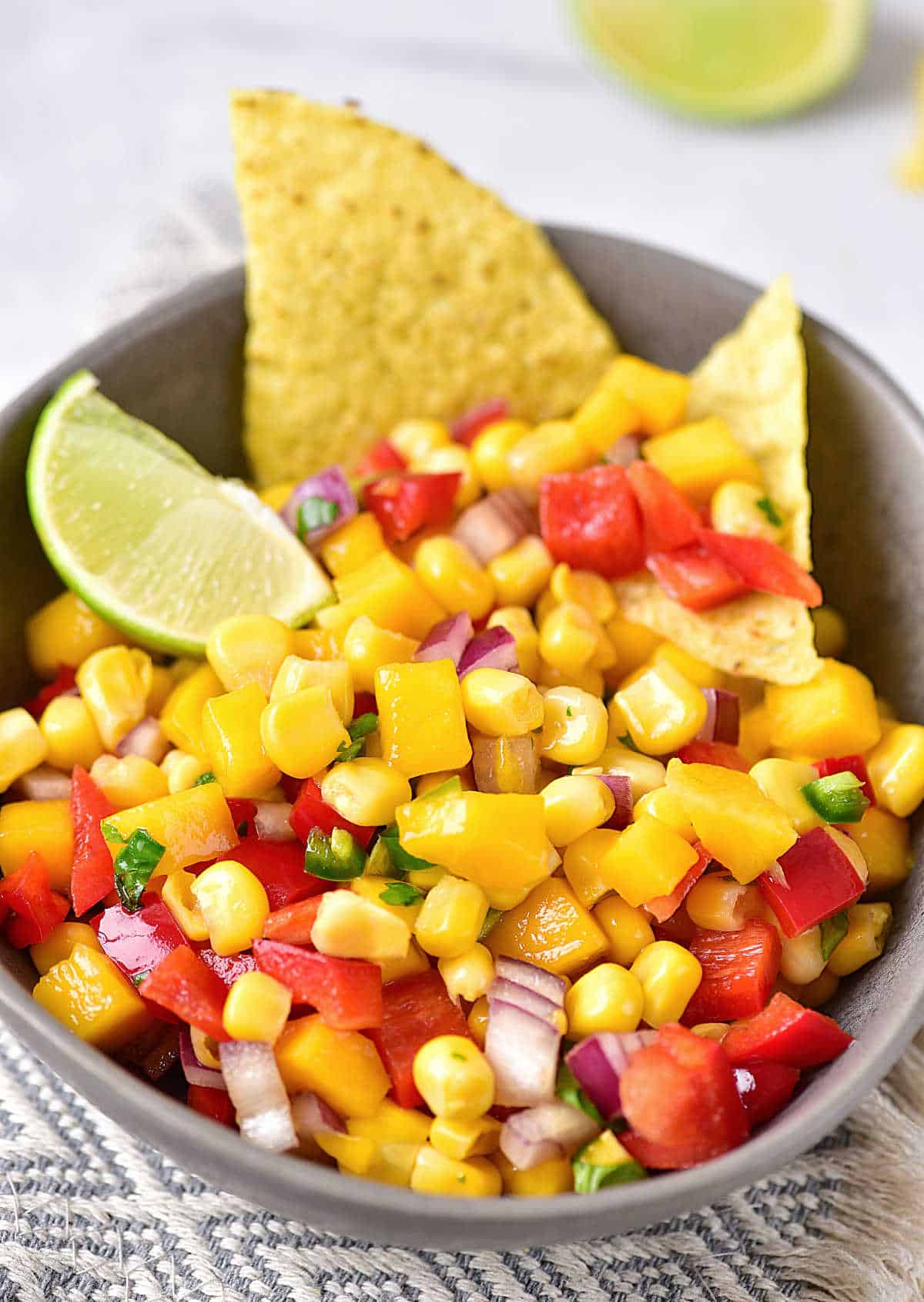 Patterned grey white cloth with grey bowl on top containing corn mango salsa with chips and lime wedge.