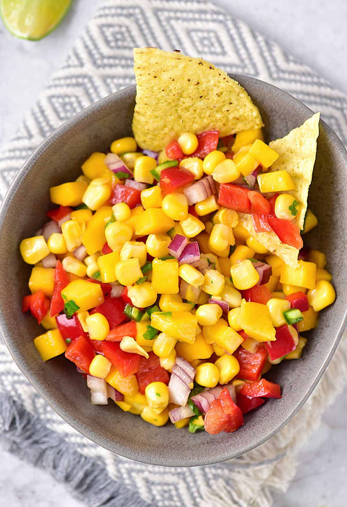Top view of grey bowl with mango corn salsa with chips. Patterned grey and white cloth underneath. 