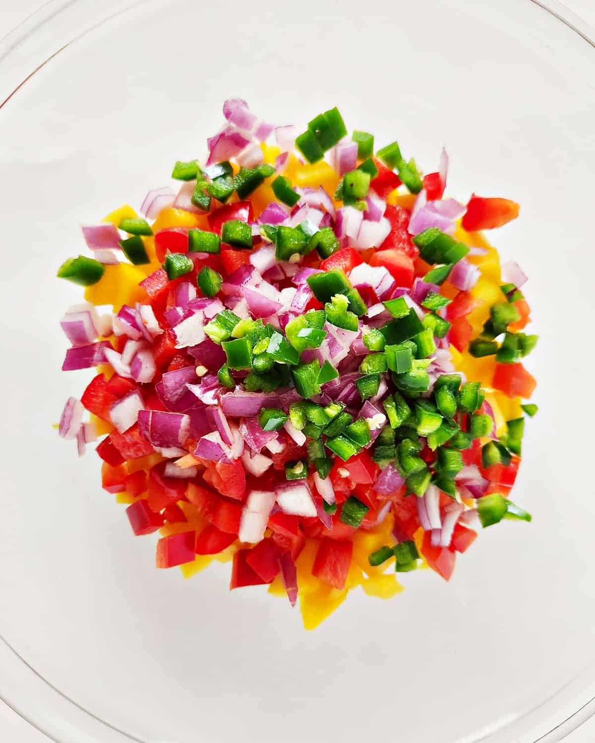 Chopped ingredients for mango peach salsa in a glass bowl on a white surface.