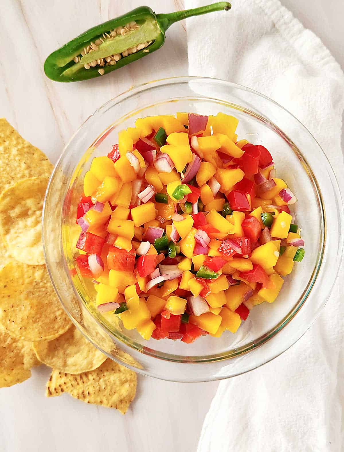 Top view of peach mango salsa in a glass bowl, white cloth, corn chips, half jalapeno pepper. White surface.
