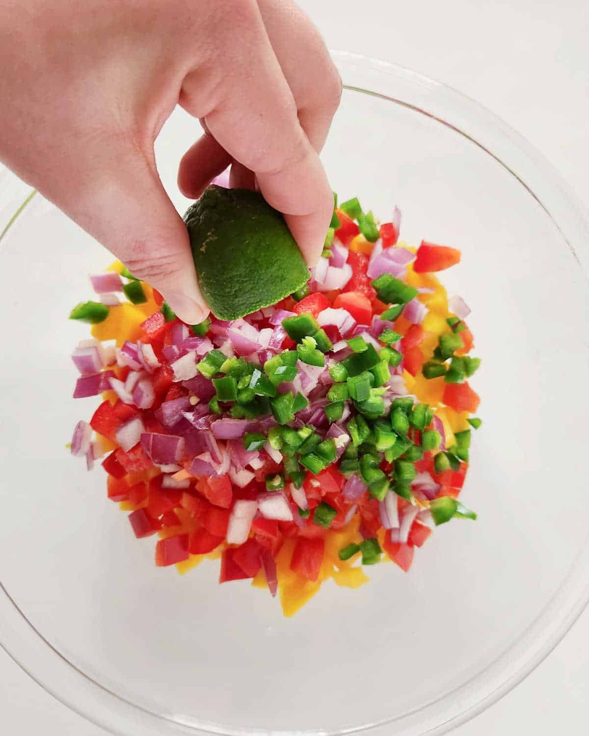 Squeezing lime juice over chopped ingredients for mango peach salsa in a glass bowl on a white surface.