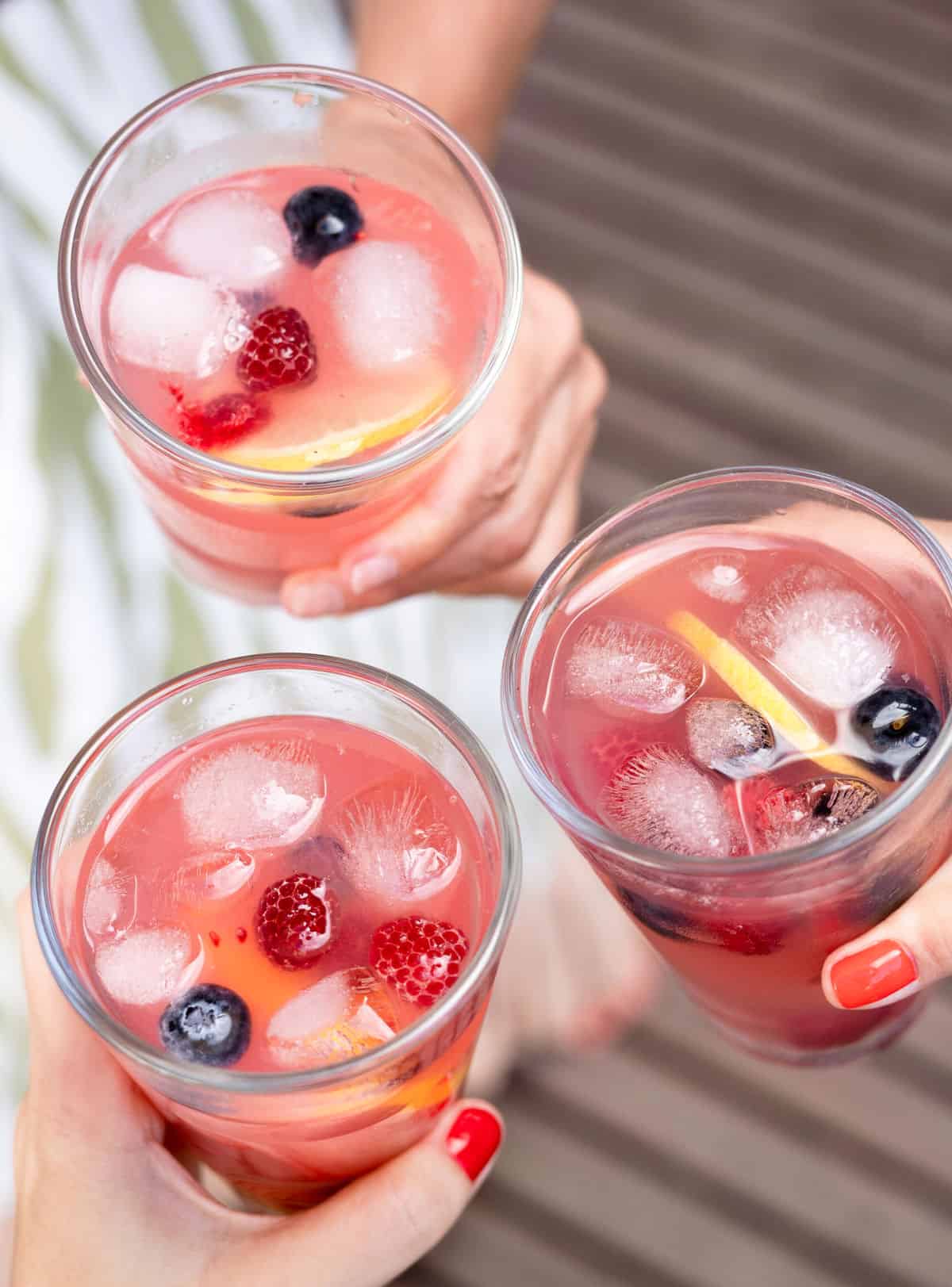 Top view of hands holding three glasses with pink lemonade, ice, and berries. 